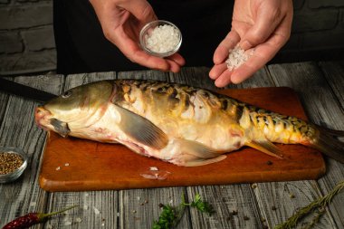 Birisi bir elinde bir kase deniz tuzu tutuyor ve yeni temizlenmiş bir balığın üzerine tuz serpiyor. Tahta bir kesme tahtasına. Yakınlarda taze bitkiler ve baharatlar görülebilir..