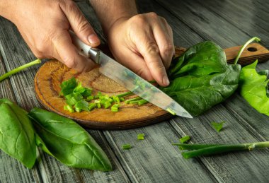 İki el ustalıkla yeşil soğan ve yapraklı yeşillikleri parlak, kırsal bir mutfakta tahta kesme tahtasında doğruyor. Malzemelerin canlı renkleri vurgulanmış..