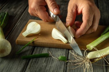 Elleri ahşap kesme tahtasındaki yeşil soğanları ustalıkla doğruyor, yakınlarda sebzeler, sıcak bir mutfakta aşçılık hazırlığı anı sergiliyor..