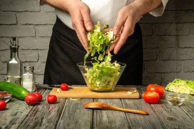 Bir şef taze sebzelerle sebzeleri ahşap bir yüzeyde cam bir kaseye karıştırır. Etrafını saran parlak domatesler ve salatalıklar sağlıklı bir yemek hazırlıyor..