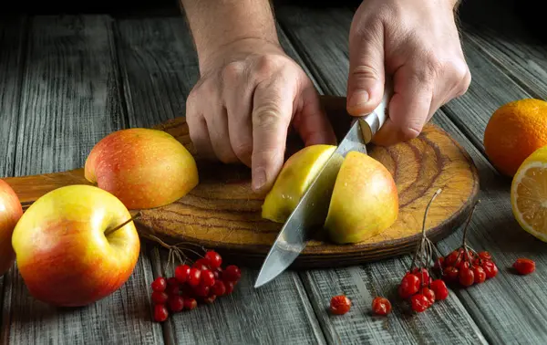 Eller tahtadan taze elmalar kesmekle meşgul. Dekor, portakal ve kırmızı böğürtlen gibi çeşitli meyveleri içererek, canlı bir mutfak atmosferi yaratıyor..