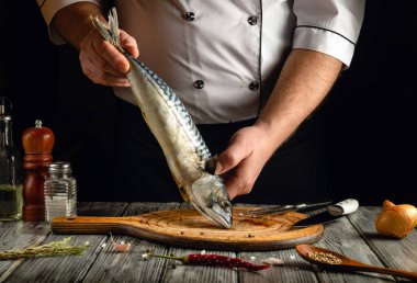 Bir şef sıcak bir mutfakta tahta bir kesme tahtasının üzerinde taze bir uskumru tutar. Hazırlık alanını çevreleyen çeşitli baharatlar, otlar ve malzemeler aşçılık atmosferini artırıyor..