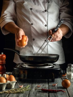Bir şef, fırının üstünde tavaya taze yumurta kırarak yemek hazırlıyor. Kır mutfağı çeşitli yemek malzemeleriyle dolu. Mutfak yaratıcılığını ön plana çıkarıyor..