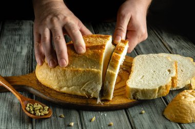 Elleri ustalıkla dilimle sıcak, taze pişmiş ekmeği tahta bir tahtada dilimle. Mutfak sıcak bir atmosfer yayıyor. Etrafa saçılmış kakule kapsülleri, rahatlatıcı bir mutfak deneyimi yaratıyor..