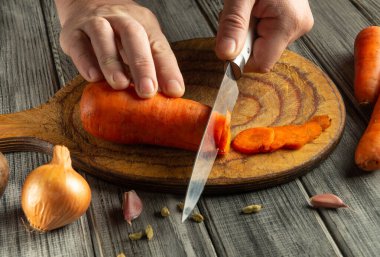 Eller, parlak turuncu bir havucu ahşap bir kesme tahtasına ustalıkla kesti. Yakınlarda soğan ve karanfil var. Mutfaktaki taze malzemelerin altını çiziyorum..