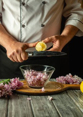 Yetenekli bir şef kırsal bir mutfakta çalışırken akasya çiçekleriyle dolu bir kasenin üzerinde bir limon dilimler. Dekor ahşap elementler ve yumuşak doğal aydınlatma içeriyor.