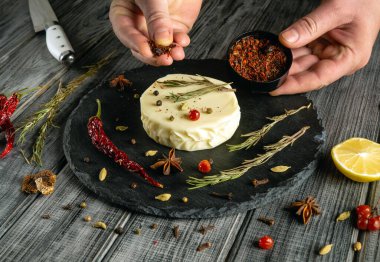 Bir şef, taze otlar, kırmızı biberler ve limon dilimleriyle kaplanmış kurutulmuş otlar ve baharatlarla dolu bir peynir turunu dikkatlice mevsimlendirir..