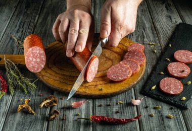 Bir şef, ahşap bir kesme tahtasının üzerinde bir sosis kütüğünü ince kurşunlara ustalıkla keser. Kurutulmuş otlar ve baharatlar tahtanın etrafını sararak mutfak atmosferini güçlendiriyor..