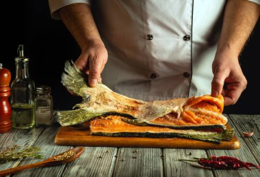 Bir şef ustaca fileto yapıyor taze somon, canlı renkler ve dokular sergiliyor. Kırsal bir ahşap yüzeyde çeşitli otlar ve baharatlar sıralanarak mutfak atmosferi oluşturulur..