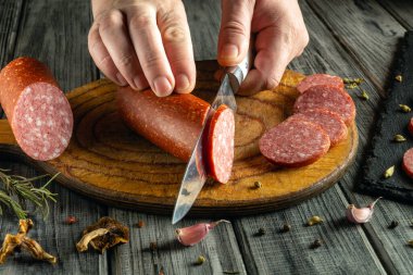 Eller ustalıkla salamı tahta bir tahtada dilimlere ayırdı. Çevreleyen malzemeler arasında otlar, sarımsak ve baharatlar yer alıyor..