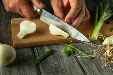 Taze taze soğanların arasında, ahşap kesme tahtasındaki beyaz soğanları ustalıkla dilimleyen eller, canlı bir mutfak atmosferi sergiliyor. Kır mutfağı yemek pişirmek için davet edici bir zemin sağlar..