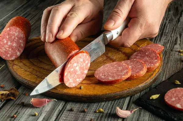 Bir insan ahşap kesme tahtasıyla salamı ustalıkla ince dilimlere ayırır. Taze malzemeler ve baharatlar, sıcacık bir mutfakta canlı bir atmosfer yaratıyor..