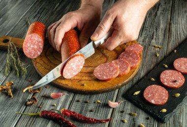 Bir insan ahşap bir tahtanın üzerinde ustalıkla sosisi dilimler, yakınlarda otlar ve baharatlar sıralanır, sıcak ve davetkar bir mutfak atmosferi yaratır..