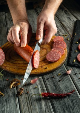 Elleri ahşap bir tahtanın üzerinde yuvarlanmış bir sosisi ustalıkla kesiyor. Çevreleyen malzemeler arasında kurutulmuş bitkiler, kırmızı biber ve sıcak bir mutfak atmosferinde bulunan soğan soğanları yer alıyor..