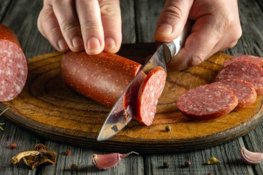 Eller, ahşap bir kesme tahtasının üzerinde duran bir sosis kütüğünü kesmek için dikkatlice bir bıçak kullanır. Mutfak seti sıcak ve davetkar, yemek hazırlamak için mükemmel..