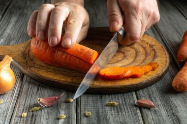 Eller ahşap bir tahtanın üzerinde taze sarımsak ve arpacık soğanları ile çevrili canlı turuncu havuçları ustalıkla dilimler. Doğal ışıklandırma kırsal mutfak atmosferini güçlendirir..