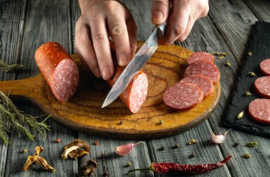 Bir insan, ahşap bir kesim tahtasının üzerinde sosis de dahil olmak üzere çeşitli et türlerini dikkatlice dilimliyor. Tahtanın etrafında taze bitkiler ve baharatlar var. Mutfak atmosferi yaratıyorlar..