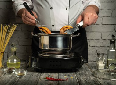 Beyaz ceketli bir şef sobanın tepesindeki bir tencereye kızarmış malzemeleri koymak için maşa ve bıçak kullanır. Mutfakta ahşap yüzeyler ve çeşitli yemek malzemeleri var..