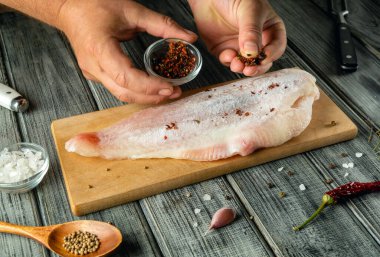 Eller taze balık filetosunun üzerine ahşap kesme tahtasına baharat serperler. Kase otlar ve tuz içerirken mutfak aletleri sıcak bir atmosferde düzgünce yerleştirilmiştir..