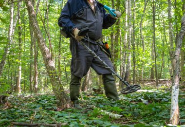 Koruyucu giysiler giyen biri, yeşil yapraklar ve ağaçlarla çevrili gür bir ormanda, ayak altında gömülü hazineleri ararken metal dedektörü kullanır..