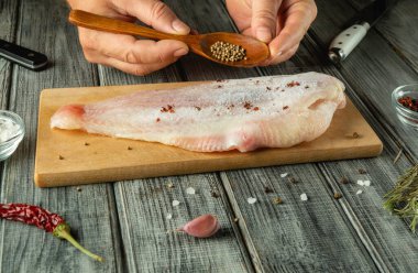 Ellerin erimiş balık filetosu üzerine kişniş serpiyor. Tahta kesme tahtasına. Çevreleyen malzemeler arasında tuz, sarımsak ve kurutulmuş biber yer alıyor..