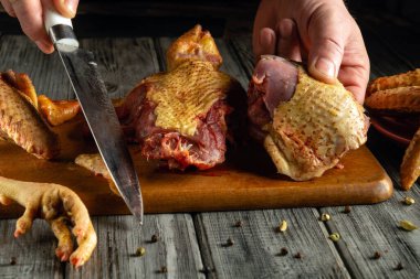 Yetenekli bir şef, tahta kesme tahtasındaki kümes hayvanlarını kesmek için keskin bir bıçak kullanır. Tahtanın etrafında çeşitli tavuk parçaları var. Kırsal bir mutfakta yemek hazırlama sanatını vurguluyorlar..