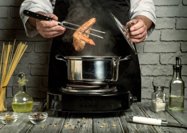 Bir şef şık bir mutfakta dikilirken buharı tüten bir sıvıyla dolu bir tencerenin üzerinde karides salıyor. Yağ, baharat ve makarna gibi malzemeler masaya yayılır..