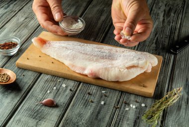 Elleri ahşap bir kesme tahtasına çiğ balık filetosunun üzerine tuz serpiyor. Yakınlarda çeşitli baharatlar ve taze otlar var, yaklaşan yemeğe lezzet katıyorlar..