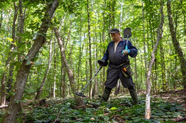 Metal detektör kullanarak canlı bir ormanda gömülü hazineleri arayan bir adam. Güneş ışığı yeşil yaprakların arasından süzülür, doğayı aydınlatır..