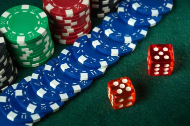 Kırmızı, yeşil ve mavi renkli kumarhane fişleri yeşil bir keçe masasında iki kırmızı zarın yanına dizilir ve heyecan ve stratejiyle dolu bir oyun gecesinin atmosferini yakalar..