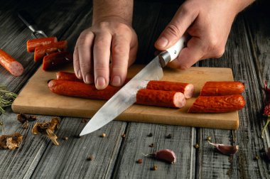 Bir kişi ahşap bir kesme tahtasındaki sosisleri kırsal bir mutfakta bıçakla dilimliyor. Baharatlar ve otlar kesim bölgesini kuşatıp sıcak bir yemek atmosferi yaratıyor..