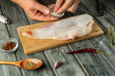 Bir insan ahşap bir kesme tahtasına uzanan balık filetosuna tuz serpiyor. Etrafı otlar ve baharatlarla çevrili. Kapalı alanda pişirme etkinliği sergiliyor..