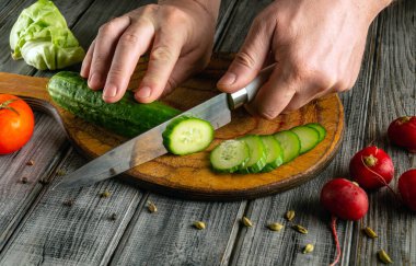 Bir insan ahşap kesme tahtasındaki salatalığı ince dilimlere ayırıyor. Yakınlarda, domates, turp ve marul mutfak düzenine doğal ışıkla aydınlatılmış renk katıyor..