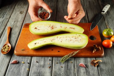 Eller ustalıkla taze bir kabağı ikiye bölerek ve baharat ekleyerek hazırlıyor. Yakınlarda, çeşitli sebzeler ve baharatlar ahşap bir kesme tahtasına dizilir..