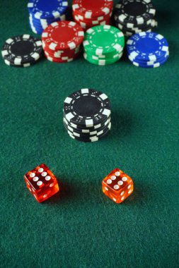 İki zar kırmızısı, bir yığın siyah poker çipinin önünde, arkalarında çeşitli renkli çipler, yeşil bir keçeli masa üzerine dizilmiş kumarhane atmosferinin tipik.