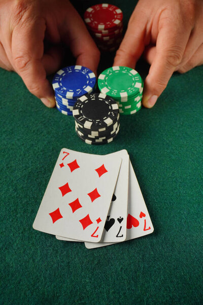 Two hands place poker chips on a green table, showcasing a pair of playing cards with diamonds and hearts in a lively casino environment at night.