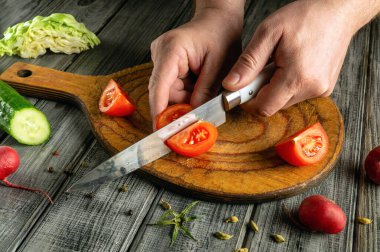 Taze domatesler kırsal bir ahşap kesme tahtasının üzerinde dilimlenir. Yakınlarda, çeşitli sebzeler bir mutfakta renkli bir hazırlık sahnesi gösteriliyor..