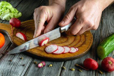 Bir insan sıcak bir mutfakta, ahşap bir tahtanın üzerinde turpları ince dilimlere ayırıyor. Yakınlarda, taze sebzeler olay yerine renk ve tazelik katar..