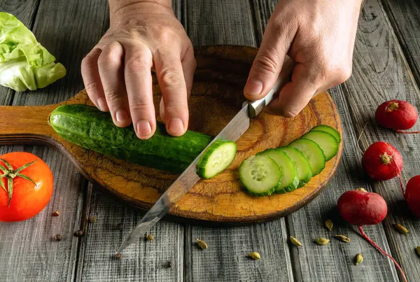 Bir çift el, yeni hasat edilmiş bir salatalığı ustalıkla yontulmuş bir tahta üzerinde keser. Yakınlarda olgun domates ve turplar mutfak çalışma alanına renk katıyor..