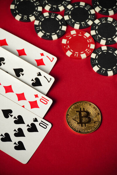 Cards depicting a pair of tens and sevens lie on a bright red table, scattered nearby are black and white poker chips and a shiny Bitcoin, symbolizing digital currency.
