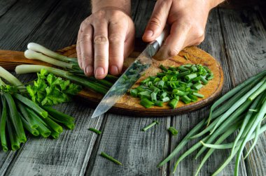Hands skillfully chop fresh green onions on a wooden cutting board. Sprigs of green onions and other herbs surround the board, highlighting a culinary preparation scene in a cozy kitchen.
