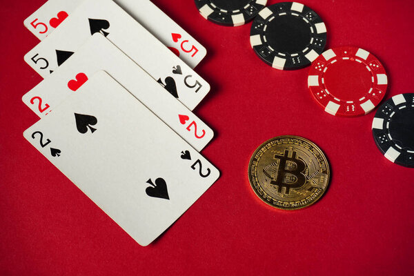 Four playing cards showing two of hearts and two of spades are arranged beside multiple poker chips and a Bitcoin coin on a vibrant red surface.
