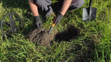 A person is digging in a grassy field, using a metal detector to find buried items. They carefully probe the soil while surrounded by tools and equipment in bright daylight