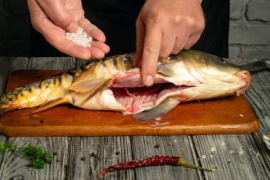 Hands expertly season a fresh fish on a wooden cutting board. Salt and a red chili pepper add flavor as the rustic kitchen enhances the culinary scene.