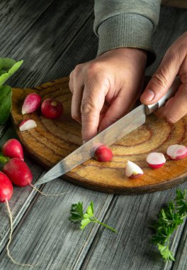 Hands skillfully cut fresh radishes on a wooden cutting board, surrounded by green herbs and other vegetables, highlighting a love for cooking and healthy eating.