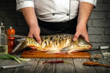 A chef handles a freshly caught fish Cyprinus carpio on a wooden cutting board in a rustic kitchen. Various spices and green onions are laid out for preparation.