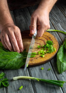 Hands are skillfully chopping green onions and leafy greens on a rustic wooden cutting board, with fresh ingredients scattered around in a cozy kitchen space.