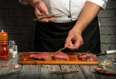 A chef is seasoning fresh beef steaks with herbs in a rustic kitchen. The wooden cutting board holds the meat, while spices and seasonings surround the scene, highlighting culinary preparation.