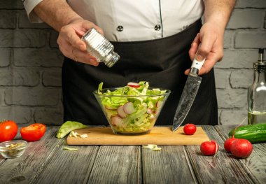 A chef in a black apron is chopping vegetables for a salad. Crisp lettuce, tomatoes, and radishes are mixed in a clear bowl. Seasoning is being added to enhance flavor.