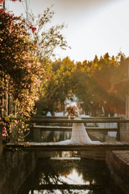 Güzel gelin gün batımında bir nehrin üzerindeki ahşap kırsal köprüde duruyor.